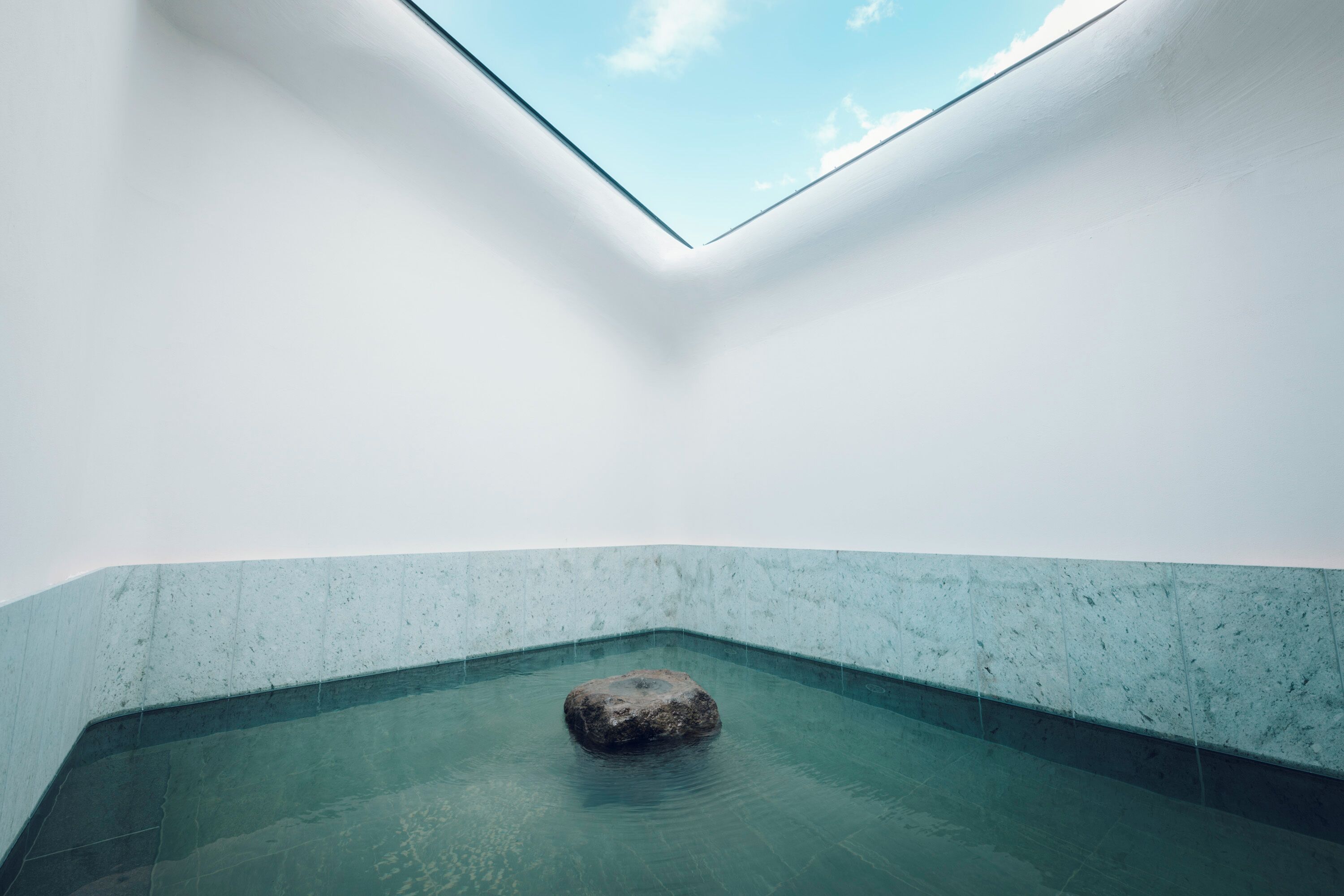 Ein minimalistischer Innenpool mit einem einzelnen Felsen, klarem Wasser und einem Oberlicht.