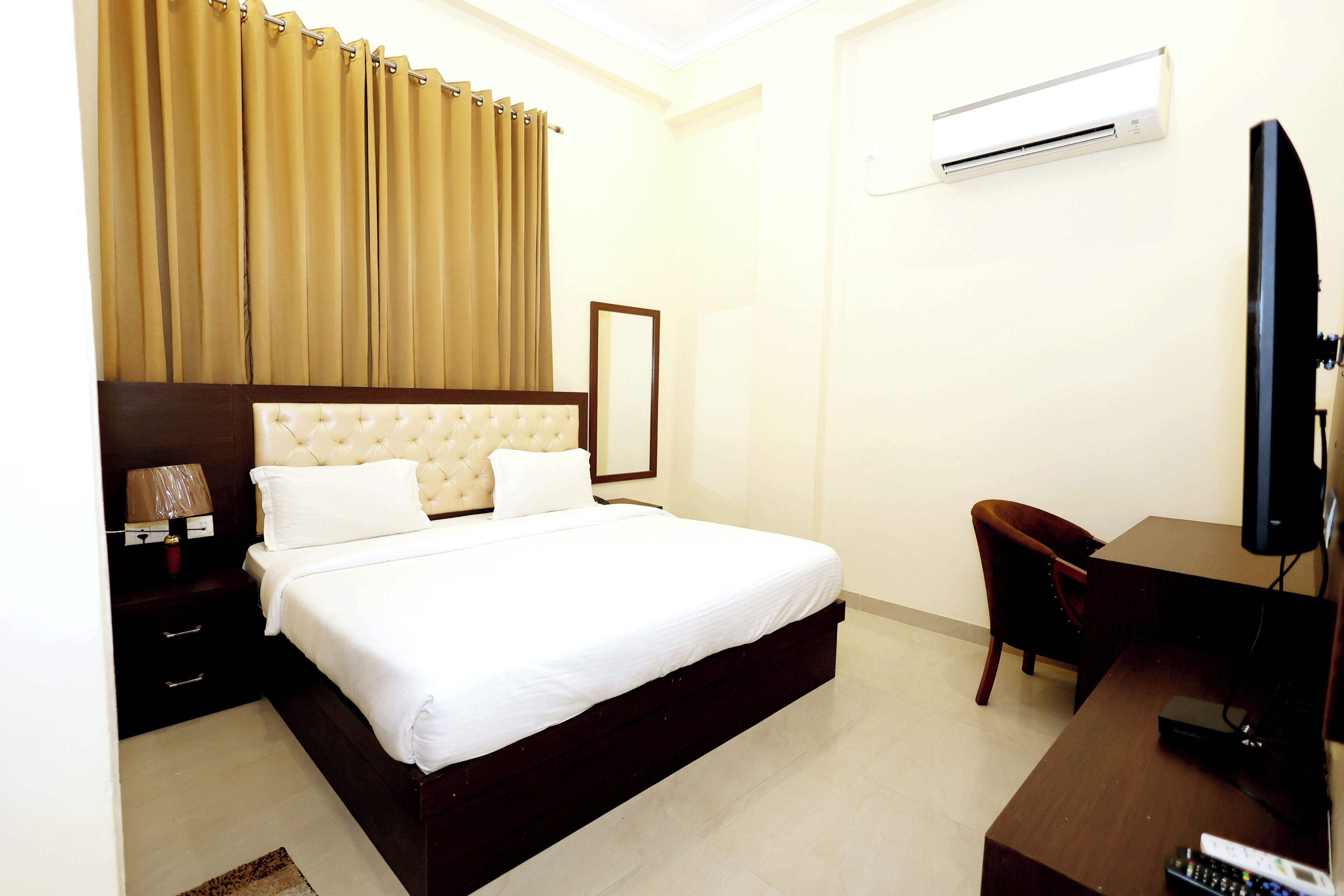 A hotel room with a large bed, a desk with a TV, a chair, an air conditioning unit, and a window with curtains.