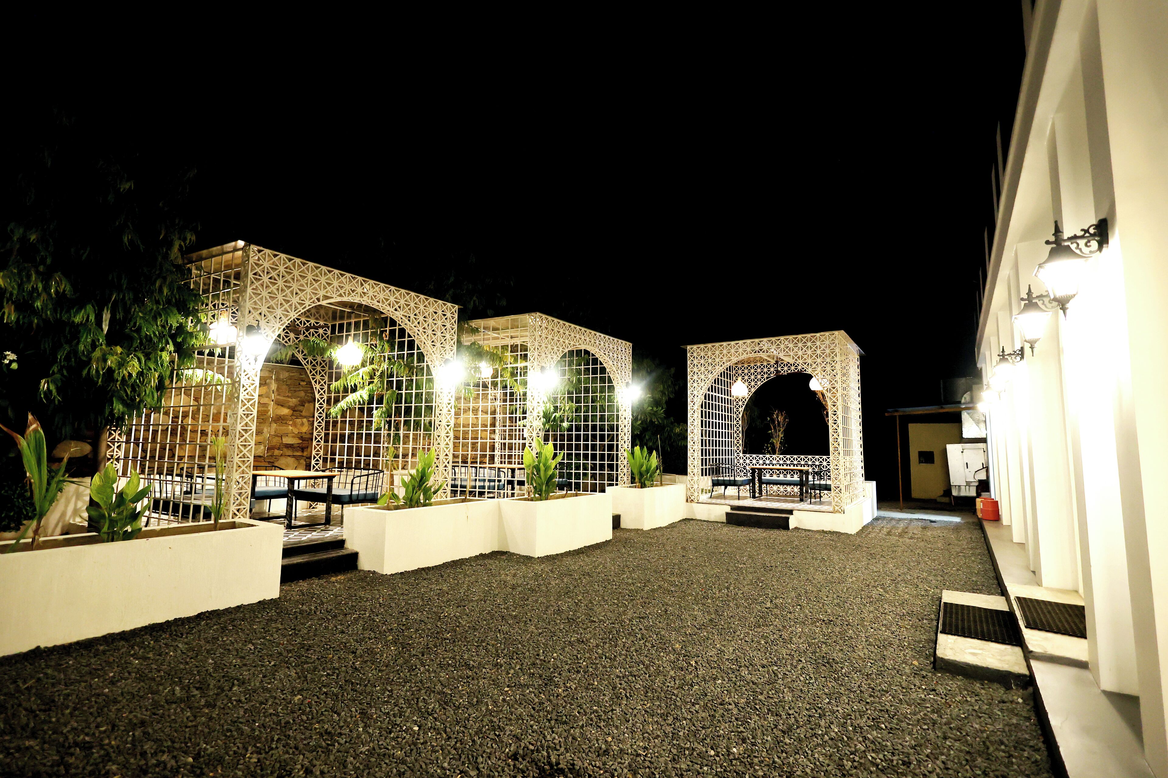 An outdoor courtyard with a series of arched structures, some with plants in white planters, and a gravel ground.