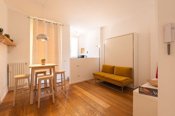 A compact living space with a wooden floor, a yellow sofa, a dining table with stools, a pendant light, and a radiator.