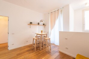 A room with a wooden floor, a table with four chairs, a small shelf with plants, and a window with curtains.