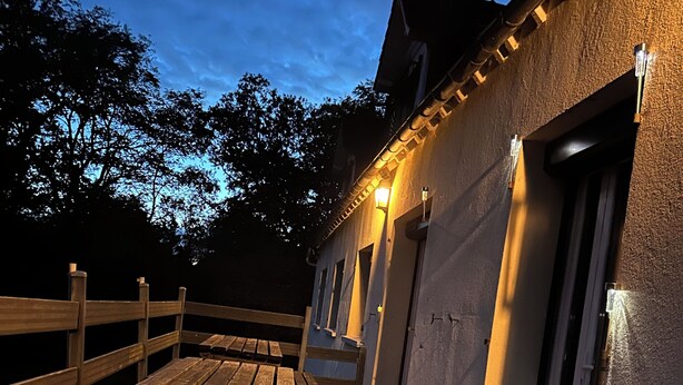 Una terrassa de fusta a l'aire lliure amb taules de picnic, un edifici amb façana rugosa i una porta amb finestre.