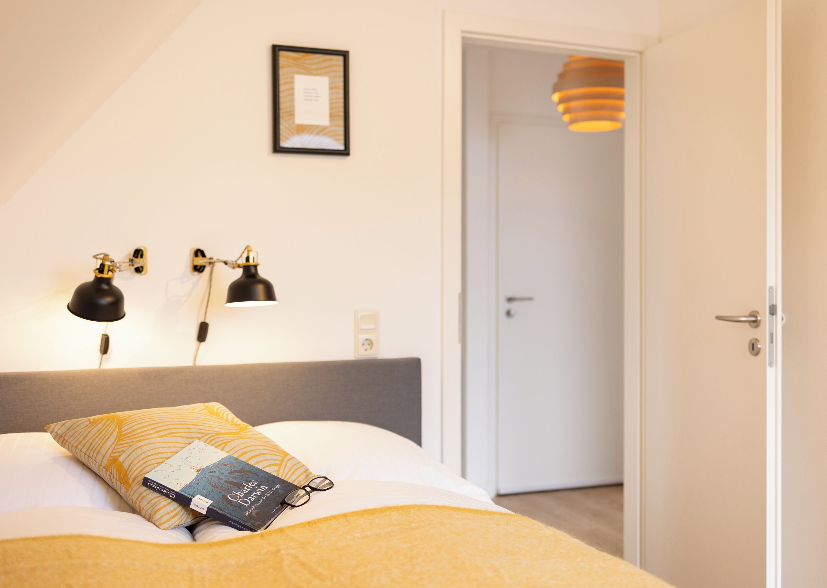 Cama bem arrumada com um livro e óculos em cima, um quadro na parede e uma porta que dá para outro quarto.