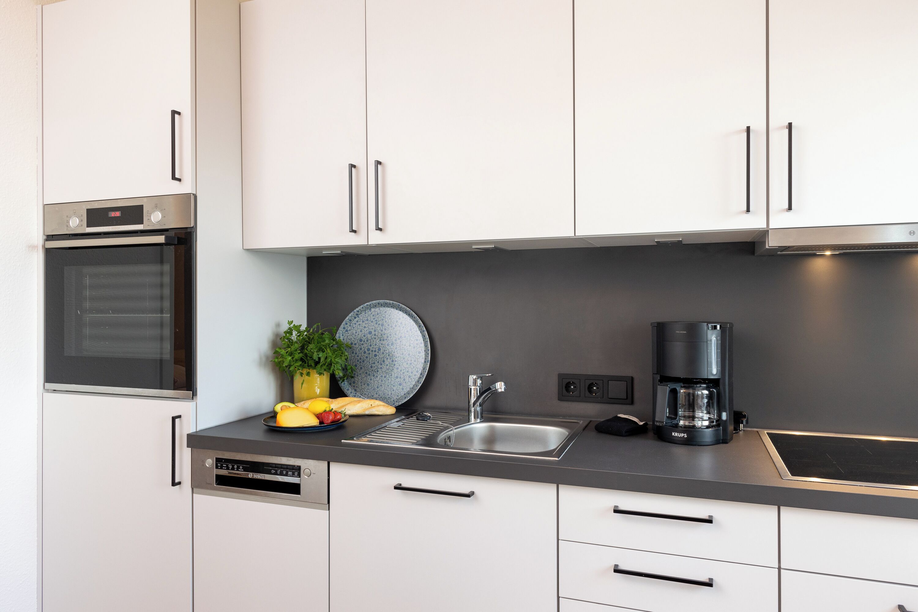Una cocina moderna con armarios blancos, horno empotrado, cafetera y lavaplatos.