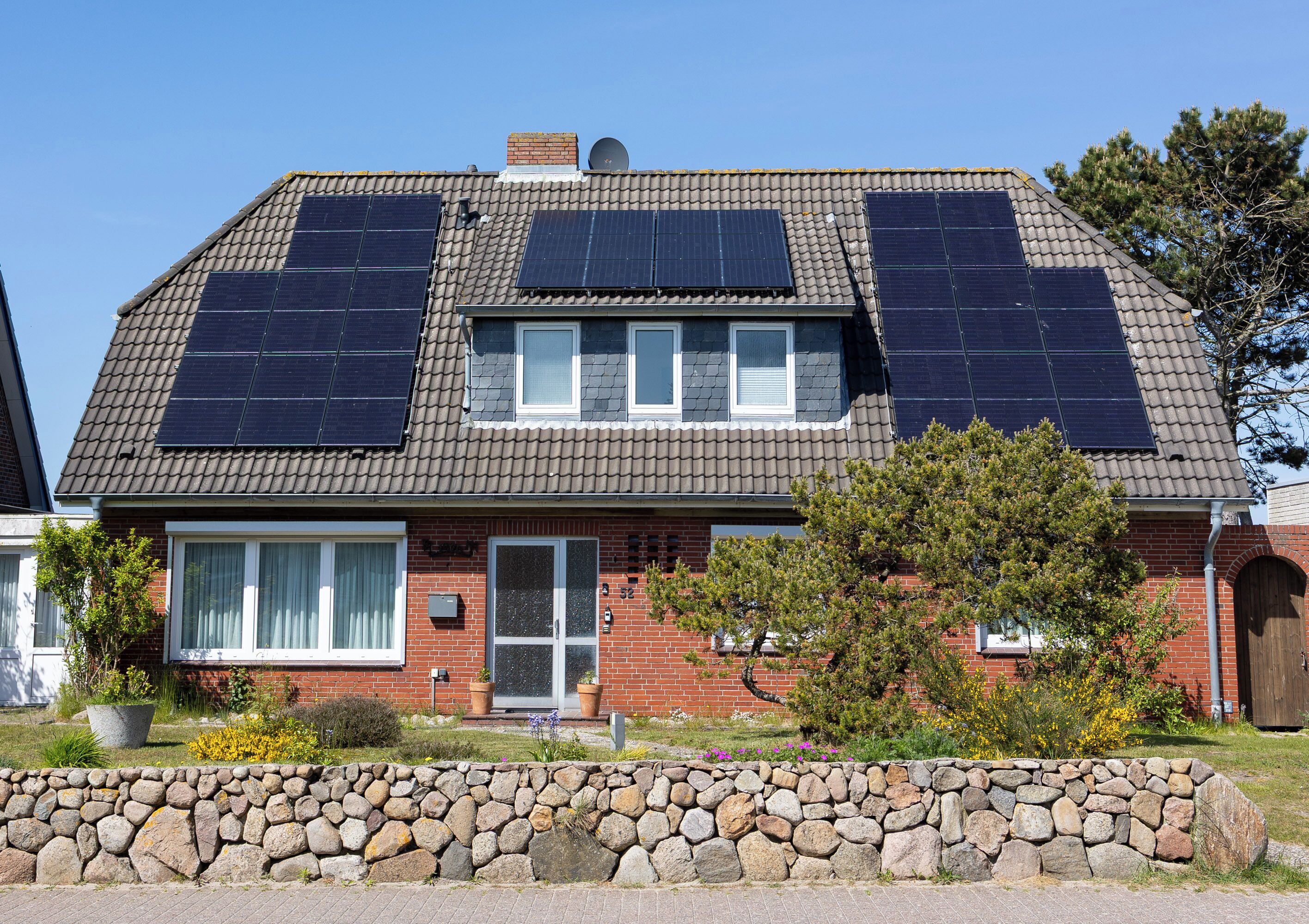 A two-story house with solar panels on the roof, a brick facade, and a stone wall in the front yard.