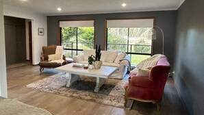 A spacious living room with a white sofa, a red armchair, and a white coffee table. There are large windows with a view of greenery outside.