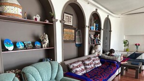 A living room with a sofa, armchairs, and a coffee table. Shelves with decorative items and books are visible.