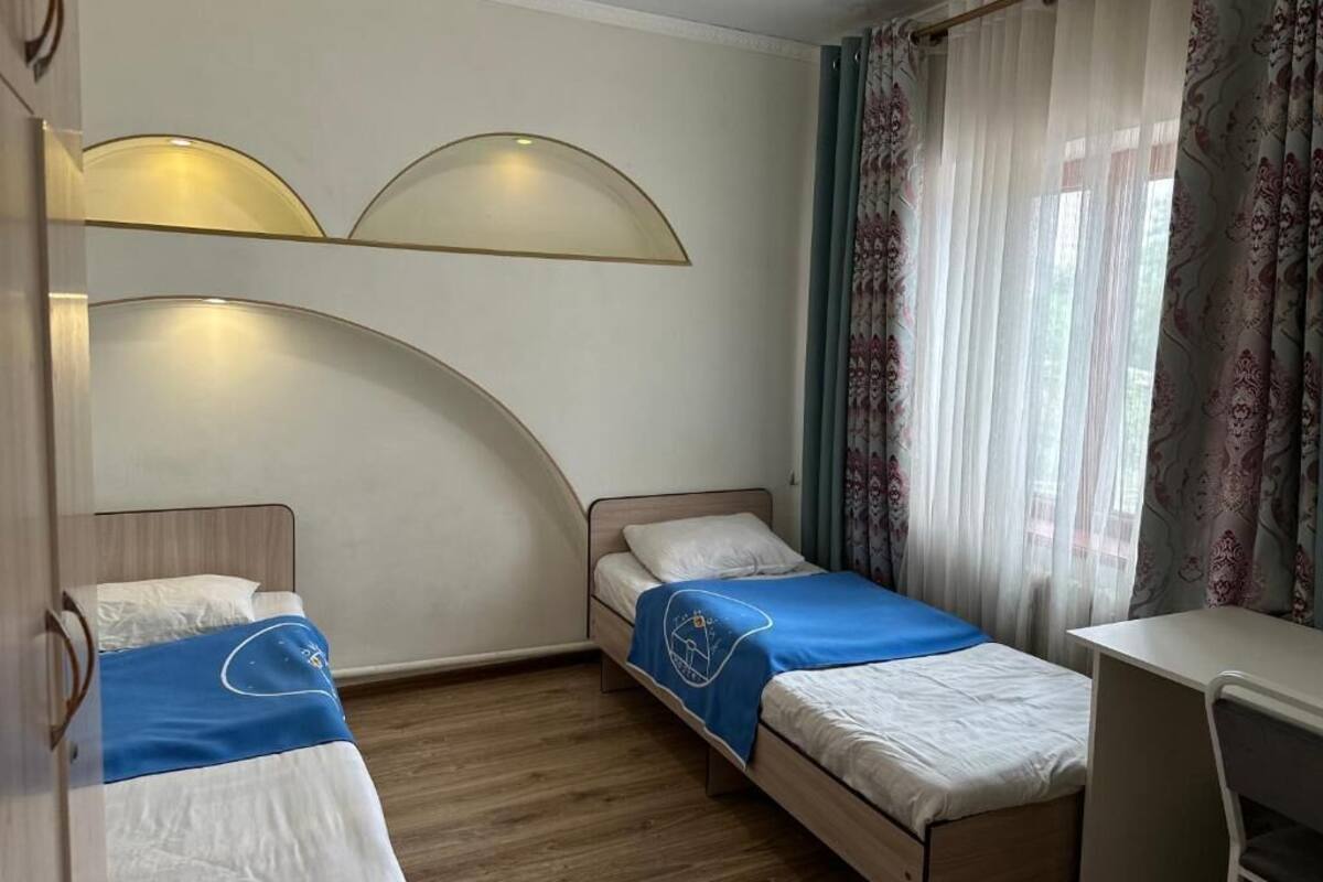 Ein kleiner, gut beleuchteter Raum mit zwei Einzelbetten, einem Schreibtisch und einem Fenster mit durchsichtigen Vorhängen.