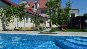 A swimming pool with clear blue water, surrounded by a house with a red roof and a covered outdoor area.