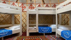 A traditional yurt with three bunk beds, each with a blue blanket featuring a logo. The interior includes a patterned rug and a wall-mounted light fixture.