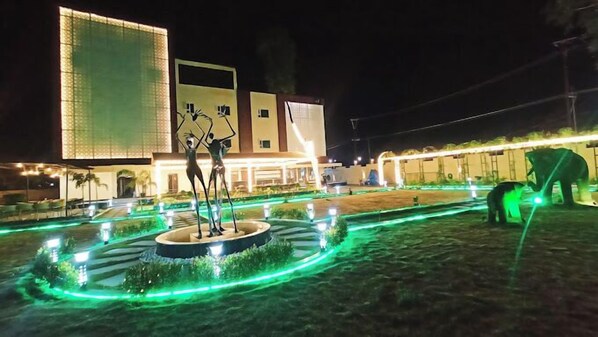 A nighttime view of a hotel courtyard with a central fountain, illuminated by green lights, and a large building with a modern design in the background.