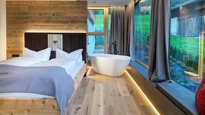 Een moderne slaapkamer met een houten hoofdeinde, een groot bed, een vrijstaand bad en een groot raam met gordijnen.