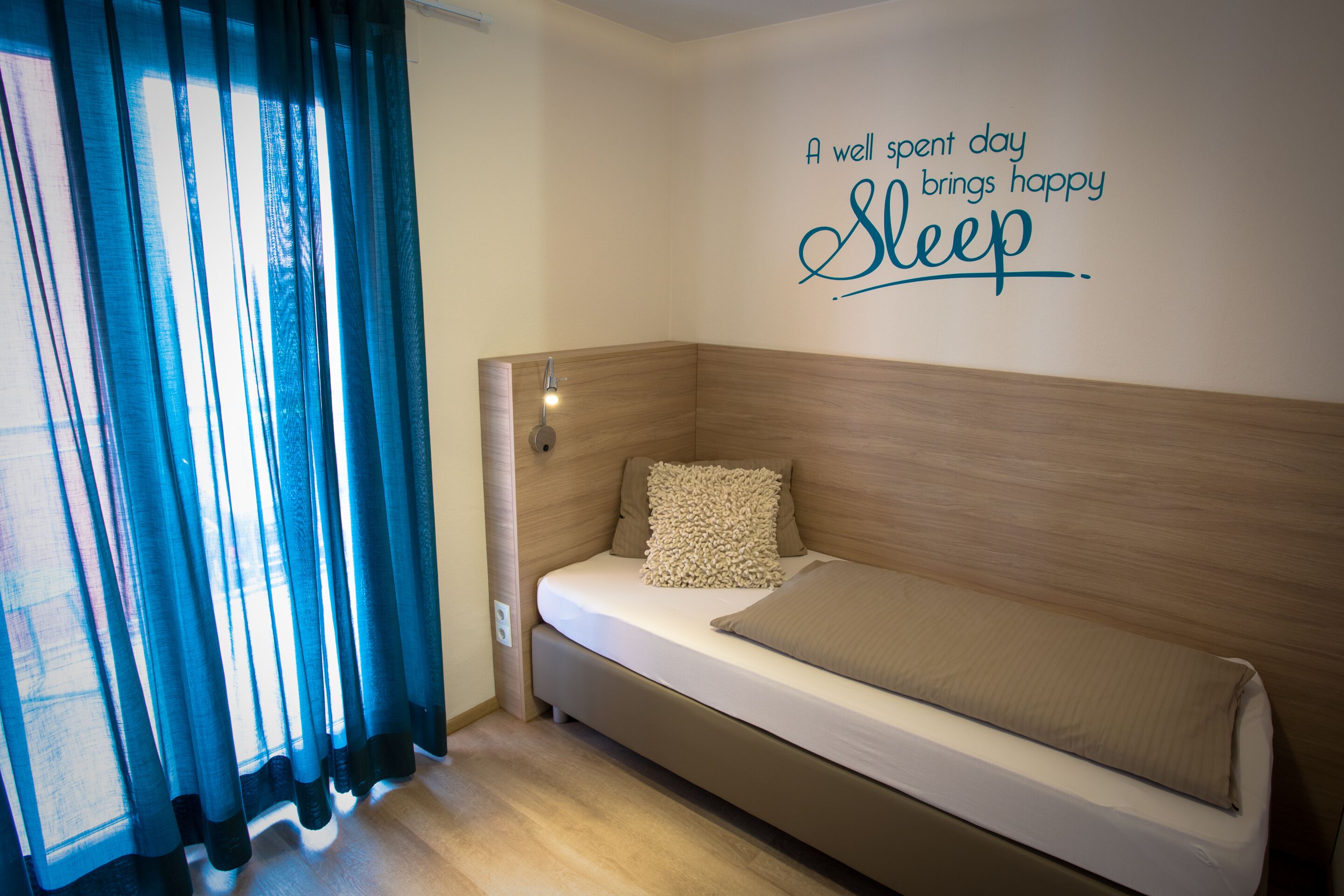 Quarto pequeno e moderno com cama, luz acoplada à parede e uma frase motivacional.