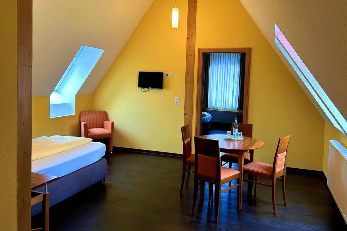 Ein kleiner, dreieckiger Raum mit einem Bett, einem Esstisch mit Stühlen, einem an der Wand befestigten Fernseher und einem Fenster mit Vorhängen.