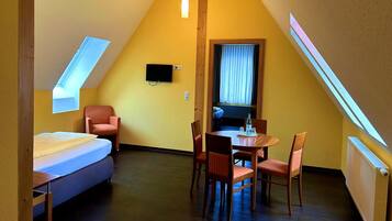 A small, triangular room with a bed, a dining table with chairs, a TV mounted on the wall, and a window with curtains.