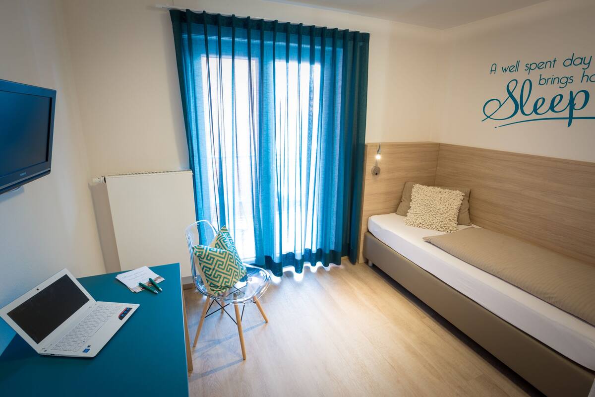 Ein Zimmer mit einem Bett, einem Schreibtisch mit einem Laptop, einem Stuhl und einem Fenster mit Vorhängen.