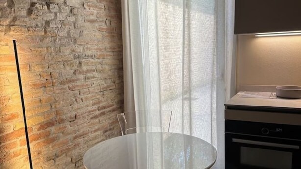 A round glass table with metal legs, a brick wall, a white curtain, and a kitchen area with a microwave. - Suite 2 (Ravenna)