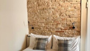 A bed with white linens and two plaid pillows against a brick wall. - Suite 2 (Ravenna)