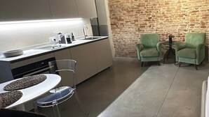 A modern kitchen with a stone wall, a dining area with a round table and chairs, and two green armchairs. - Suite 2 (Ravenna)