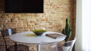 A dining area with a round table, chairs, a cactus plant, and a flat-screen TV mounted on a brick wall.
