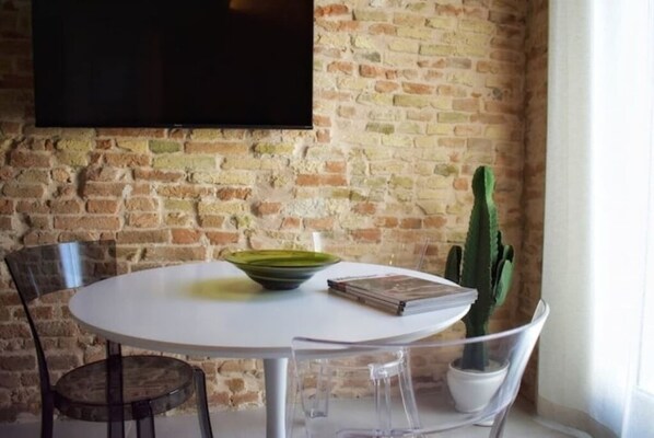 A dining area with a round table, chairs, a cactus plant, and a flat-screen TV mounted on a brick wall.