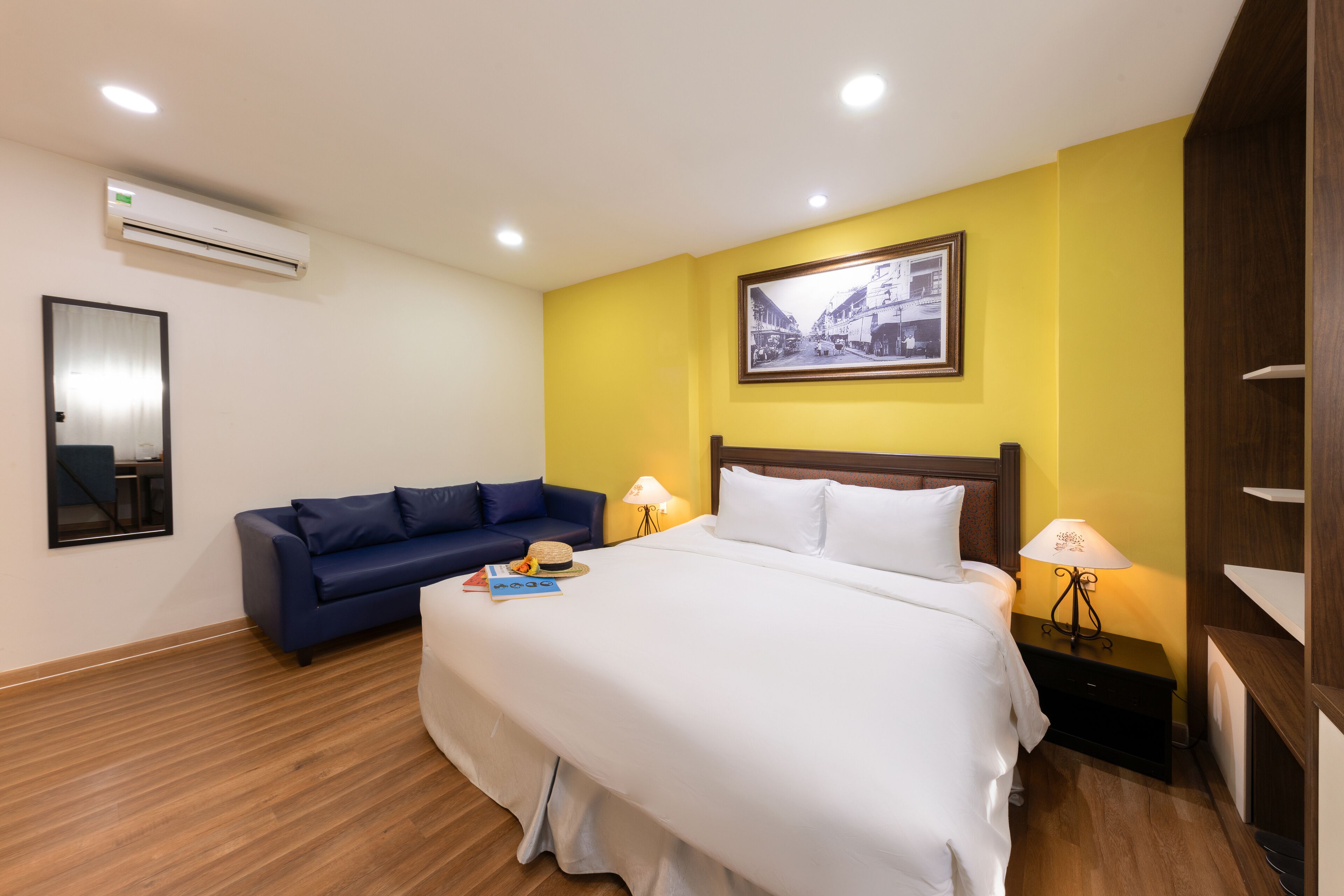 A hotel room with a bed, a blue sofa, a nightstand with a lamp, a framed picture on the wall, and an air conditioning unit.