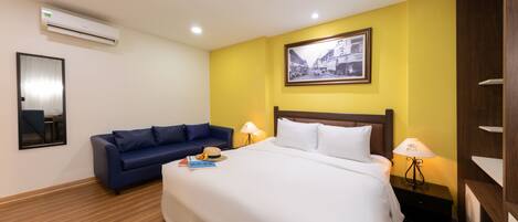 A hotel room with a bed, a blue sofa, a nightstand with a lamp, a framed picture on the wall, and an air conditioning unit.