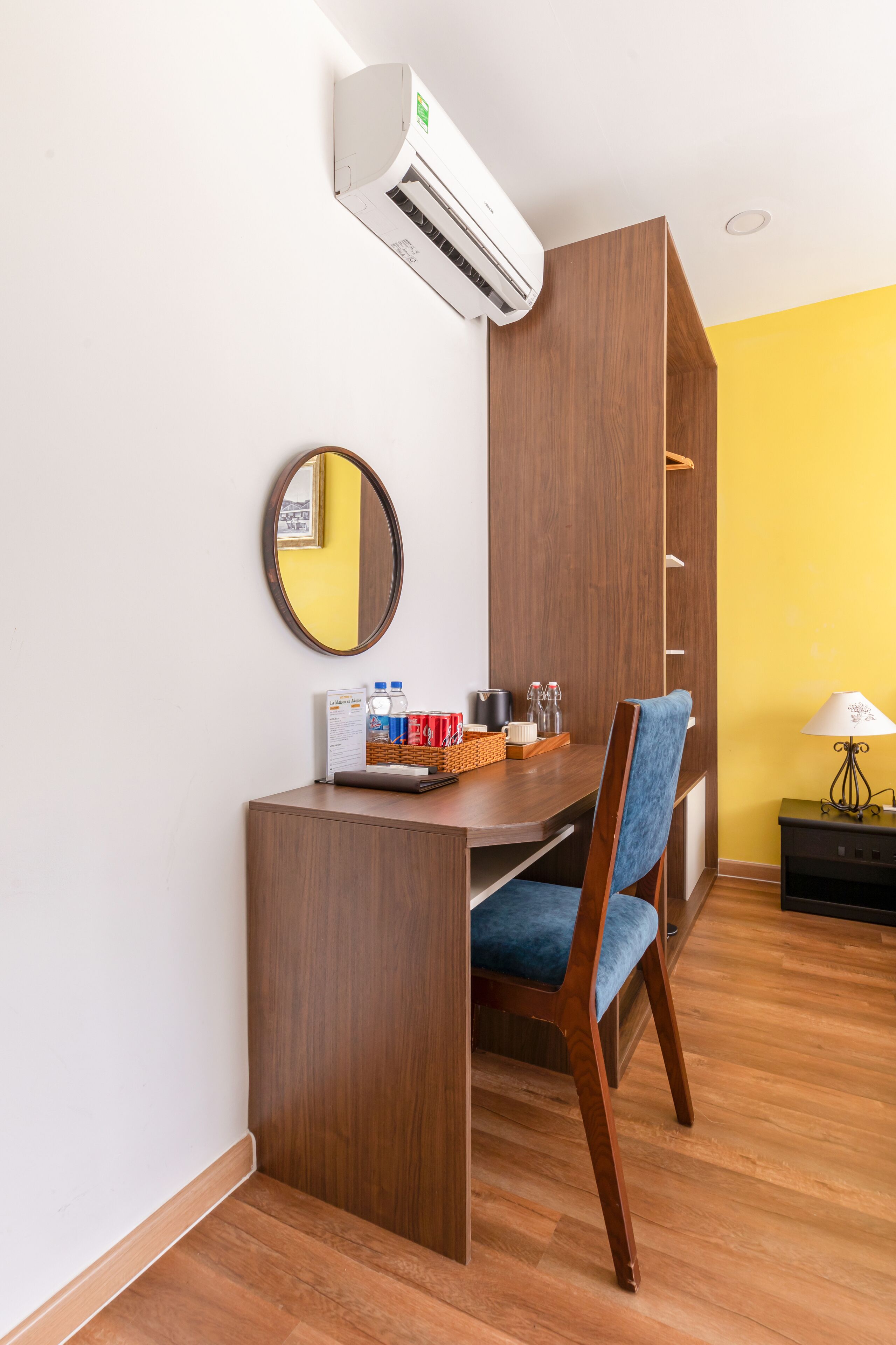 Una habitación pequeña con un escritorio de madera, una silla, una mesita con una cesta, un espejo y un aire acondicionado de pared.