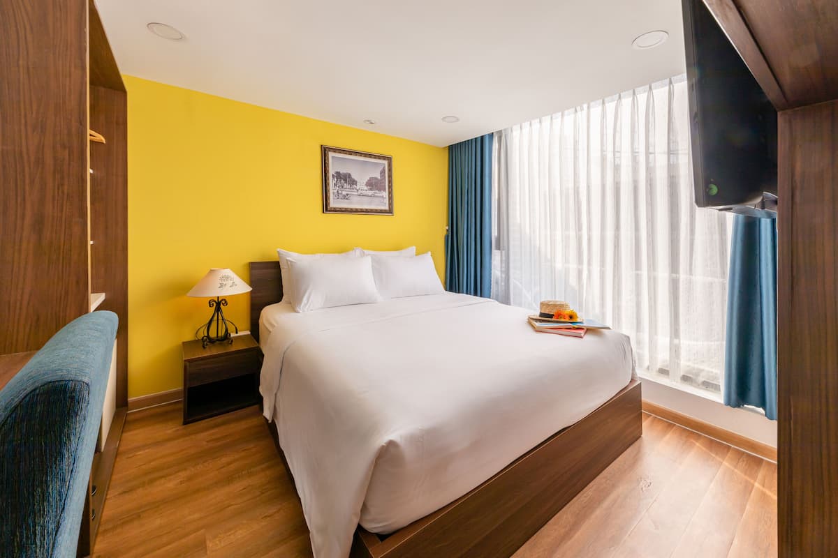 A hotel room with a large bed, wooden flooring, a nightstand with a lamp, a desk, and a TV mounted on the wall.