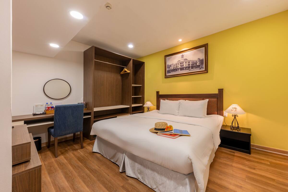 A hotel room with a bed, desk, chair, wardrobe, and a framed picture on the wall.