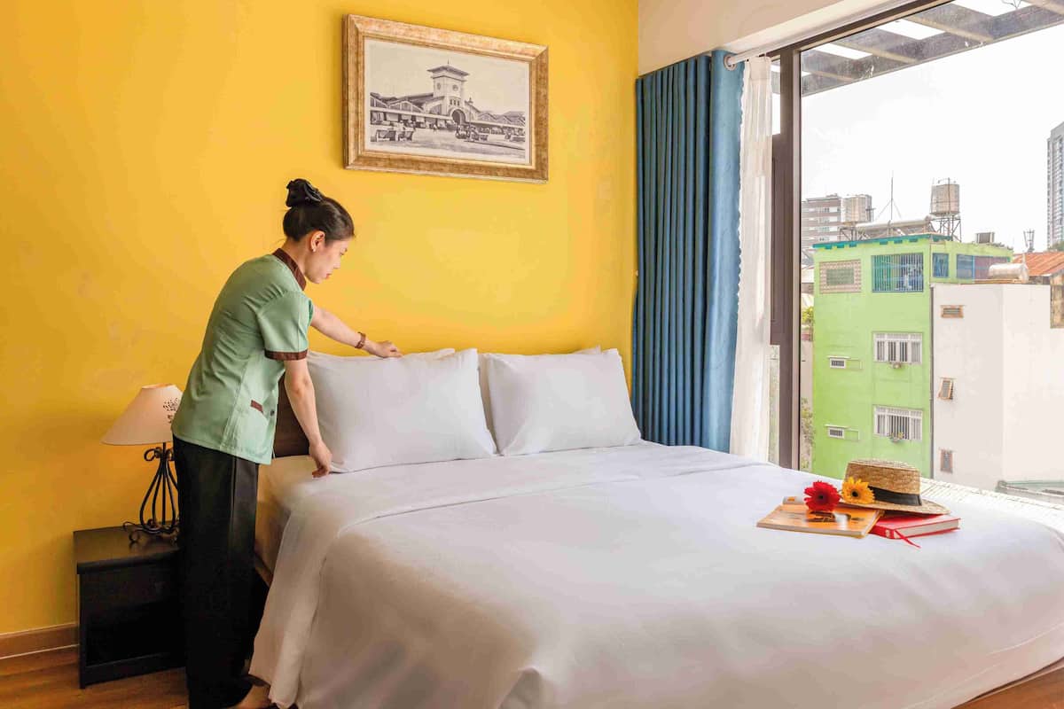 A hotel room with a bed, a nightstand, a lamp, a framed picture on the wall, and a view of buildings through the window.