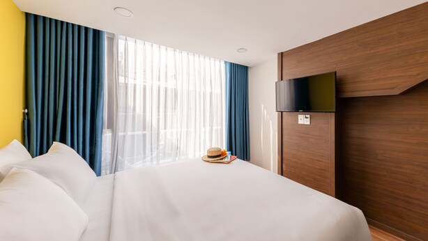 Ein Hotelzimmer mit einem großen Bett, einem an der Wand montierten Flachbildfernseher und einem Fenster mit transparenten Vorhängen.