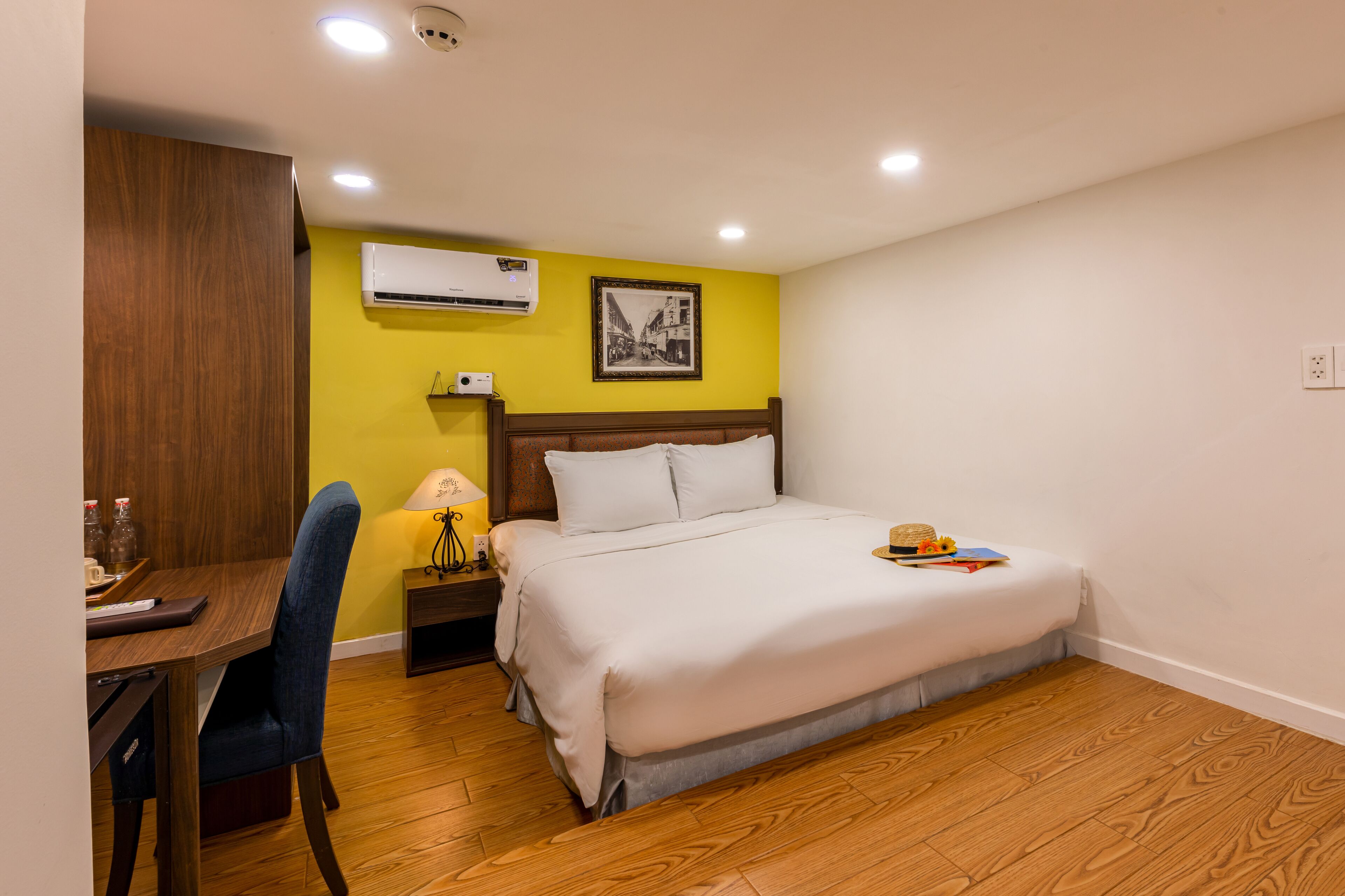 Habitación de hotel con cama, escritorio, silla, mesita de noche, aire acondicionado y un cuadro en la pared.