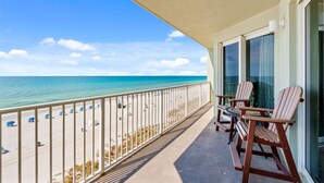 Un balcon avec vue sur la plage et l’océan. On y trouve deux chaises en bois et une table.