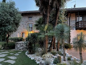 A stone building with a balcony, surrounded by a garden with various plants and a stone pathway. - Residence San Bernardo - Ap. "Monika" in the countryside, 6km from the center of Udine (Udine)