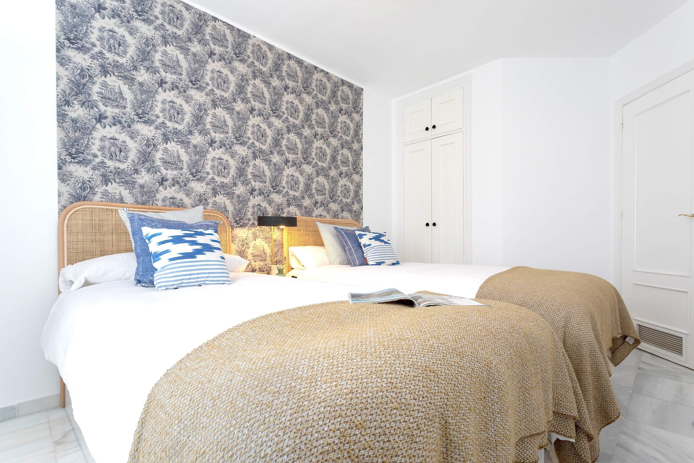 A bedroom with a large bed, two bedside tables, a nightstand, a patterned wallpaper, and a white door.