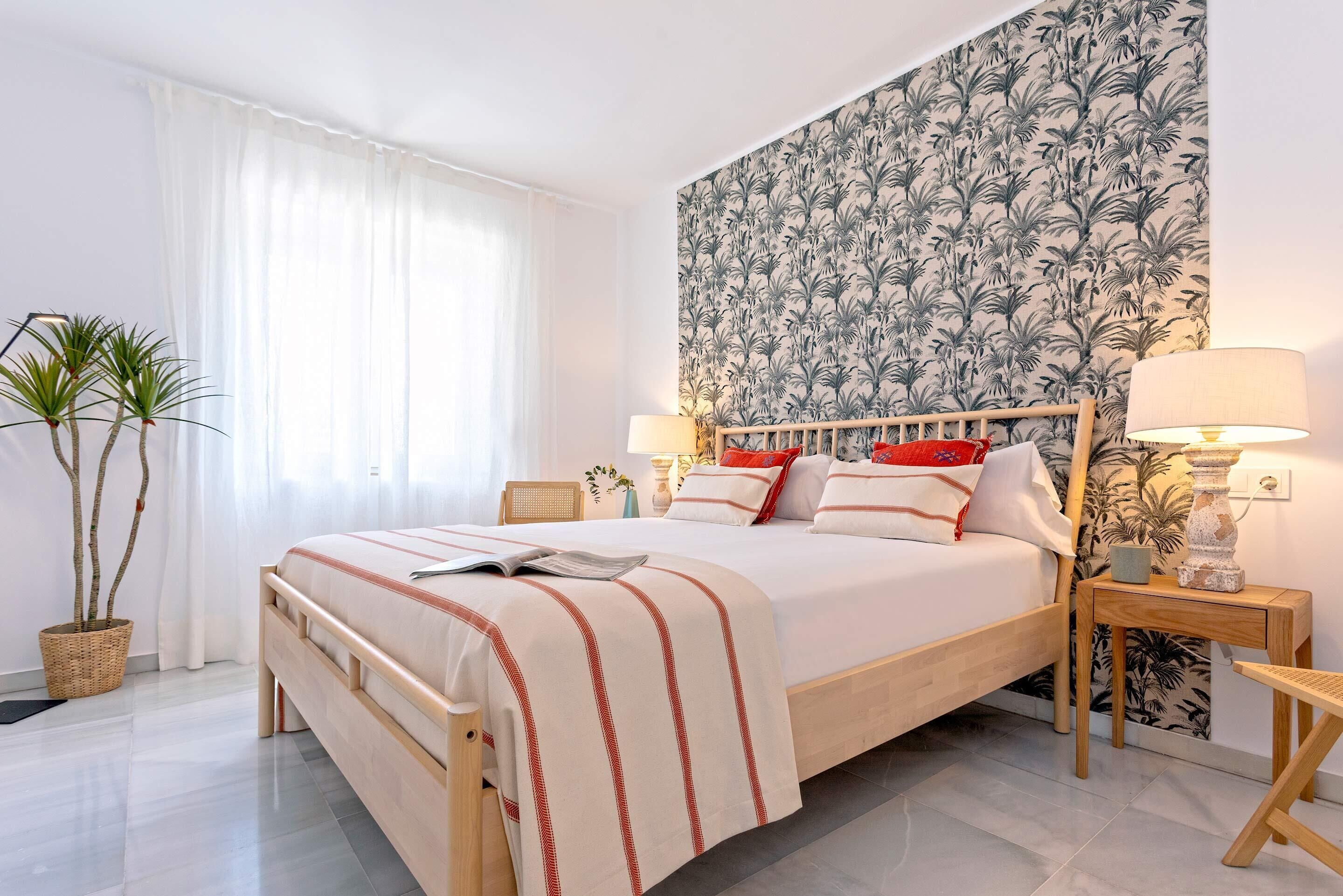 A neatly arranged bedroom with a large bed, two bedside tables, a potted plant, and a wall with a tropical pattern.