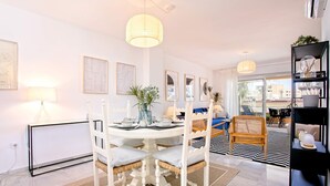 A dining area with a round table, white chairs, and a vase with flowers. There is a sofa and a black and white striped rug in the background. - Genteel Home Ricardo Soriano III (Marbella)