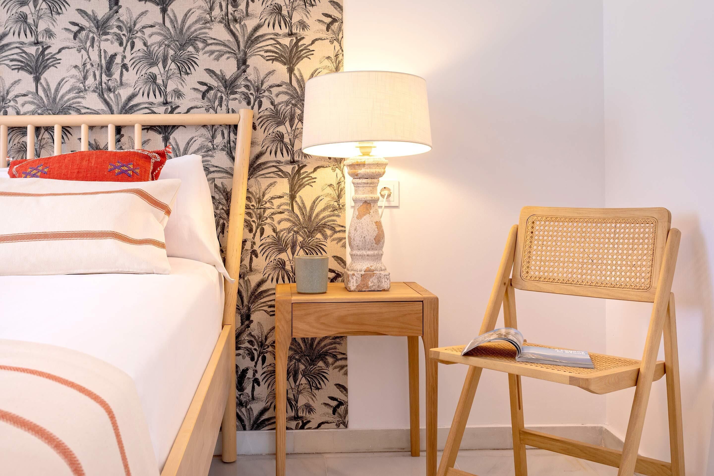 A neatly made bed with white and brown bedding, a wooden nightstand with a lamp, a wicker chair, and a wall with a tropical wallpaper pattern.