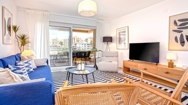 A modern living room with a blue sofa, a black and white rug, a wooden coffee table, wicker chairs, a TV, and a balcony with a table and chairs. - Genteel Home Ricardo Soriano III (Marbella)