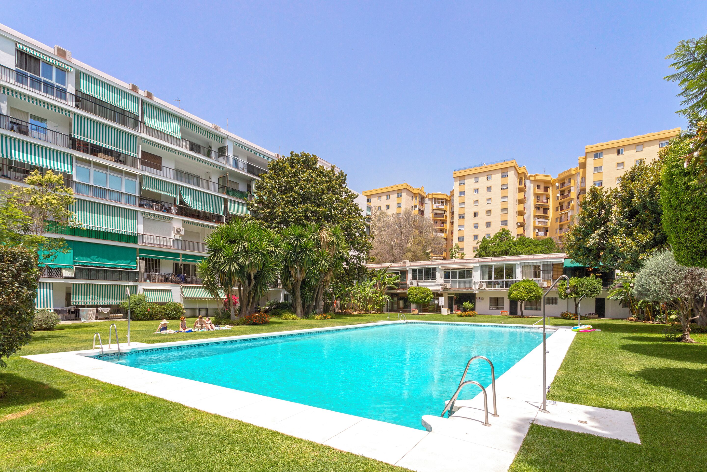 A swimming pool surrounded by a well-maintained lawn, with multi-story buildings in the background.