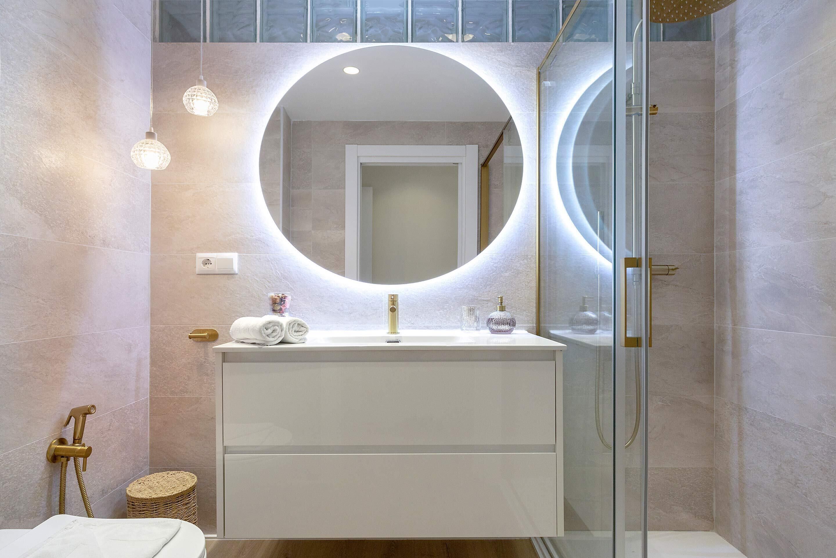 Un baño moderno con un espejo redondo, un tocador blanco, cabina de ducha de vidrio y una pequeña cesta de mimbre.