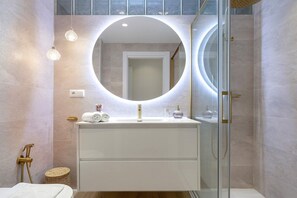 A modern bathroom with a round mirror, a white vanity, a glass shower enclosure, and a small wicker basket. - Genteel Home San Pancracio (Fuengirola)