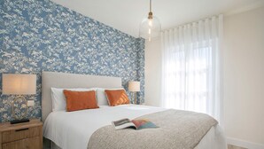 A neatly made bed with a white duvet and orange pillows, a wooden nightstand with a lamp, and a blue floral wallpaper. - Genteel Home San Pancracio (Fuengirola)