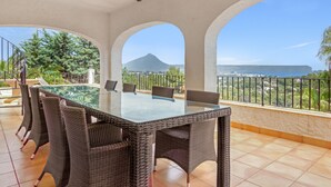 A dining area with a glass table and wicker chairs, overlooking a scenic view of a mountain and the sea. - Villa 'Casa Ann - Family Rentals' with Sea View, Private Pool and Wi-Fi (Jávea)