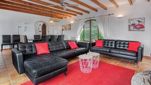 A spacious living area with a black leather sofa, red cushions, and a red rug. There are two white round tables with geometric designs. The room has wooden beams on the ceiling and a large window with white curtains. - Villa 'Casa Ann - Family Rentals' with Sea View, Private Pool and Wi-Fi (Jávea)