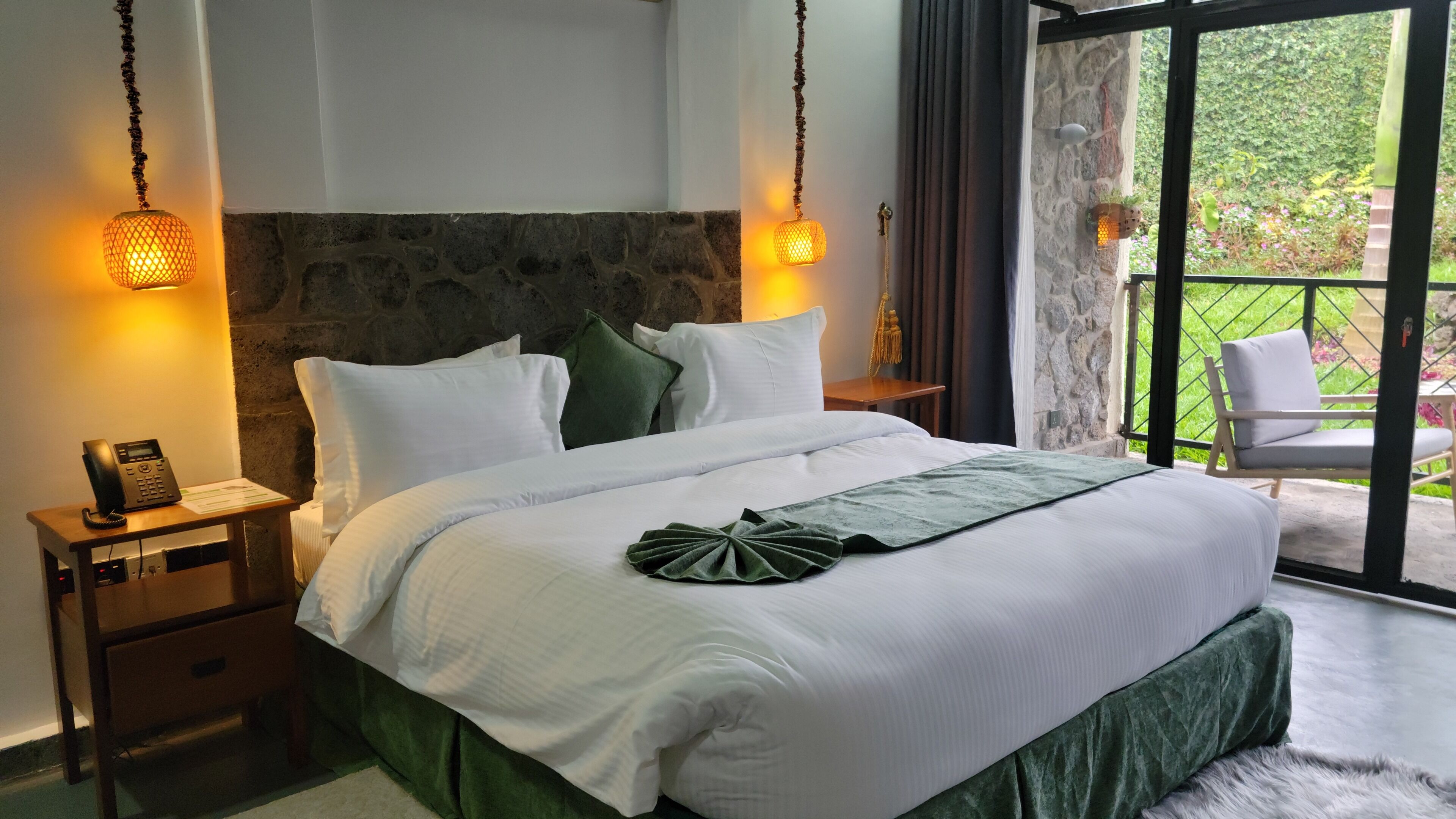 A hotel room with a large bed, two bedside lamps, a telephone on a nightstand, and a view of a garden through a glass door.