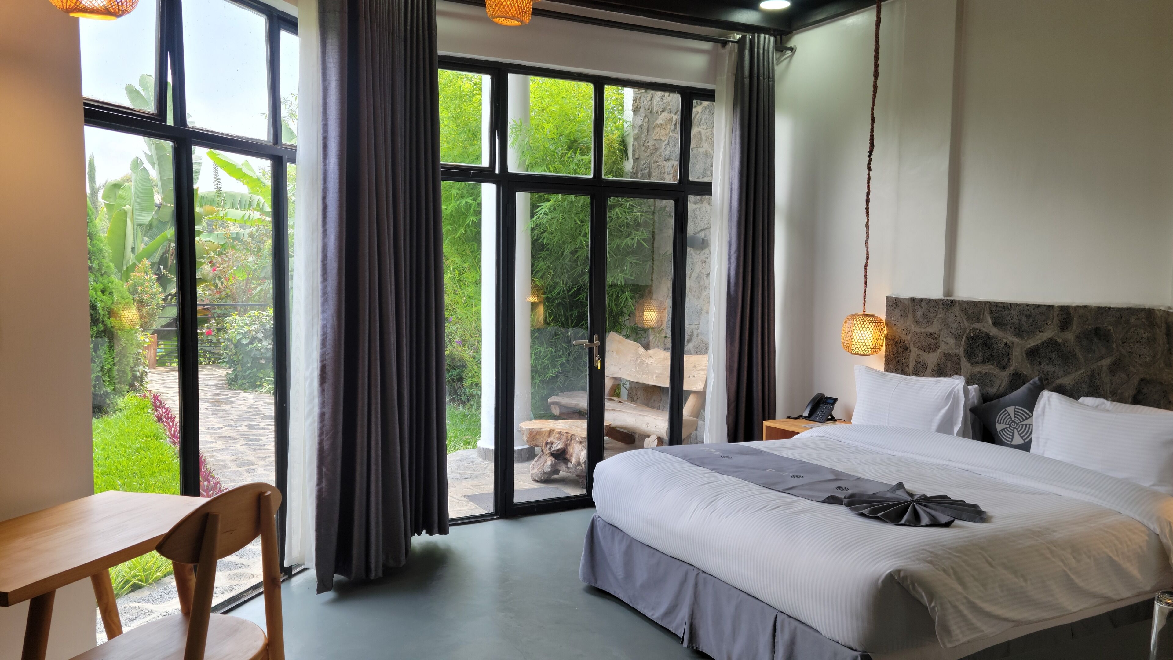 Habitación de hotel con una cama grande, una mesita de noche de madera, un escritorio y vistas a un jardín a través de amplias ventanas.