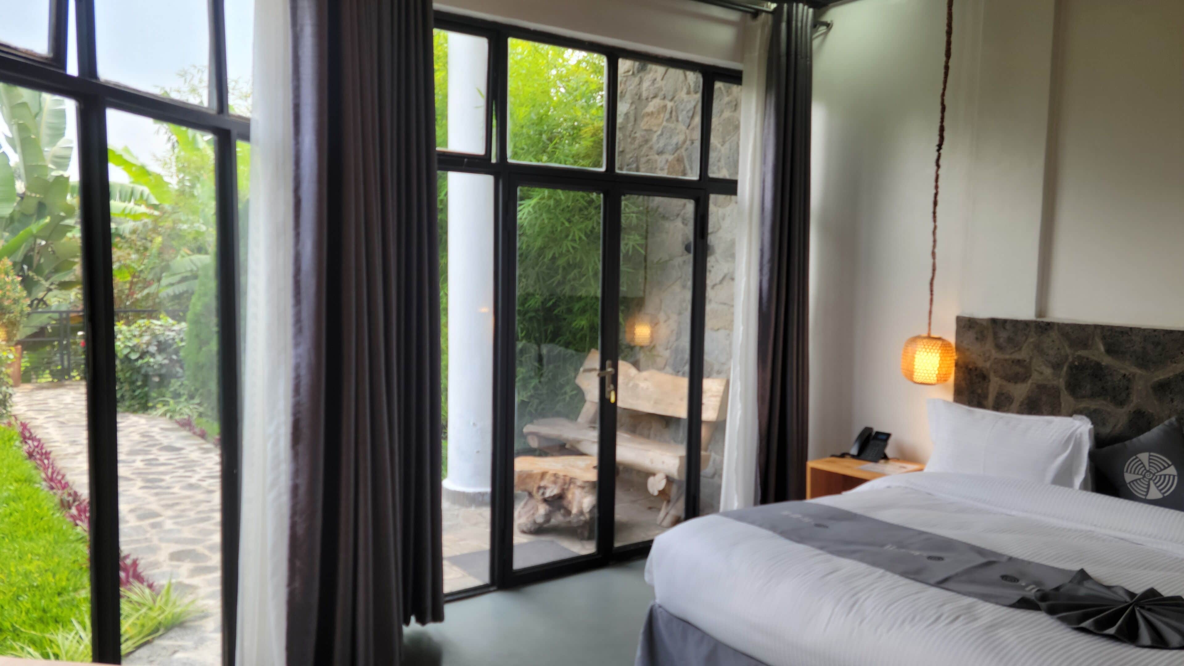 A bedroom with a large window, a bed, a nightstand, and a stone wall.