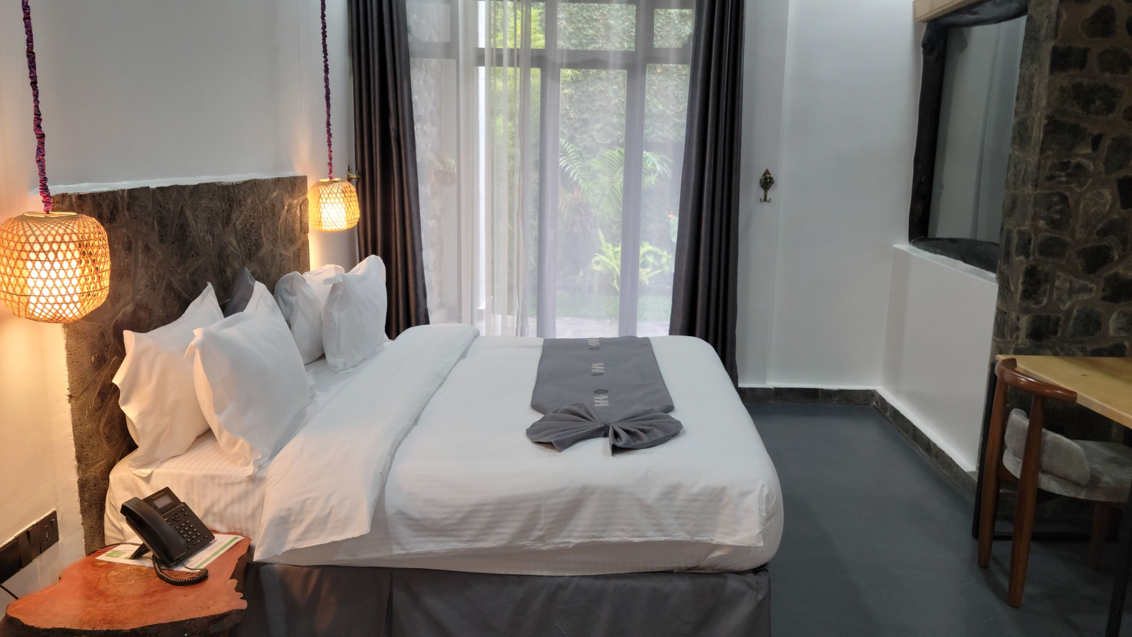 A hotel room with a bed, a bedside telephone, a lamp, a window with curtains, and a stone wall.